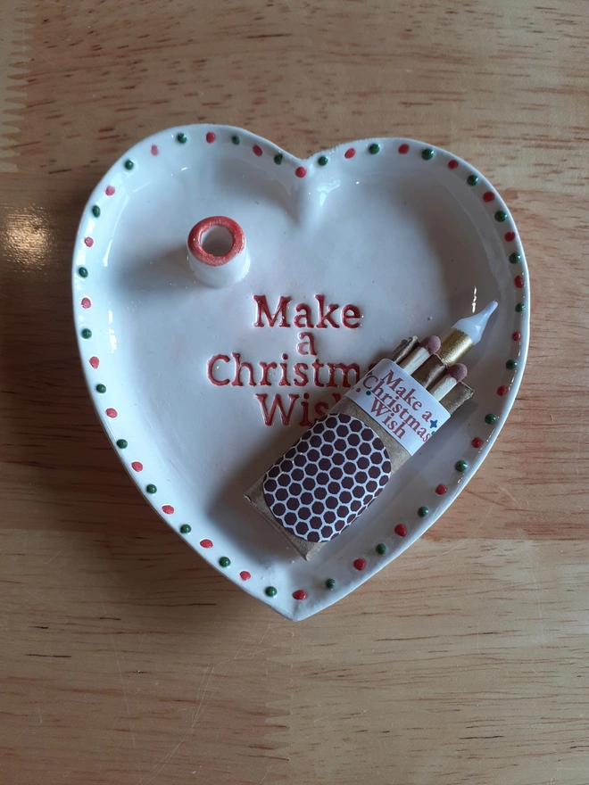 Overhead view of a handmade ceramic heart dish reading “Make a Christmas Wish”, with its coordinating match-and-candle box resting inside.