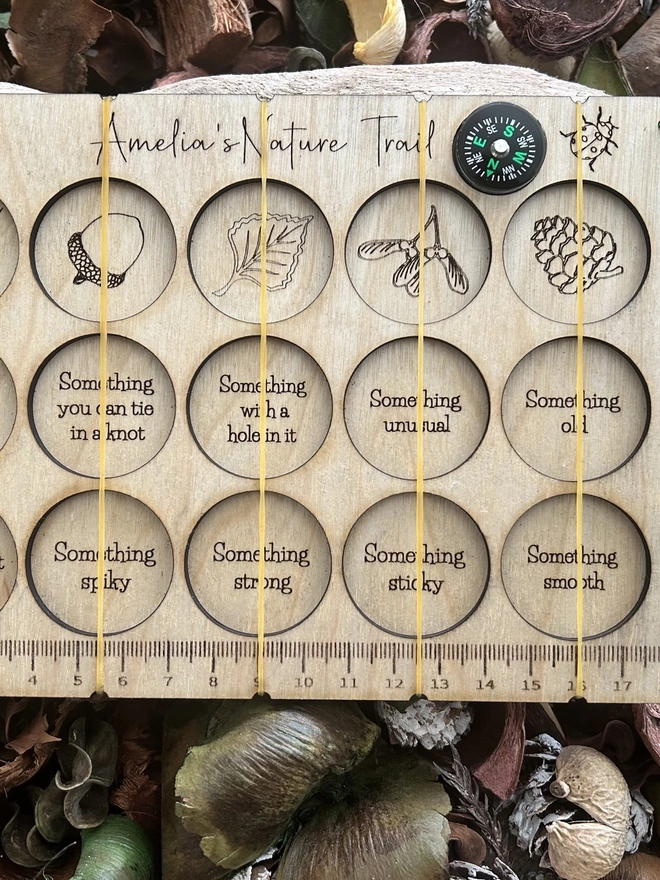 A personalised wooden nature scavenger hunt board displayed on an outdoor surface. The board is rectangular with rounded corners and engraved with 15 small collection slots held by clear bands. A custom name is engraved at the top, and a button compass with a wrist lanyard is attached for carrying. The wood has a smooth, light finish, and no items are placed in the slots; only the board is shown.