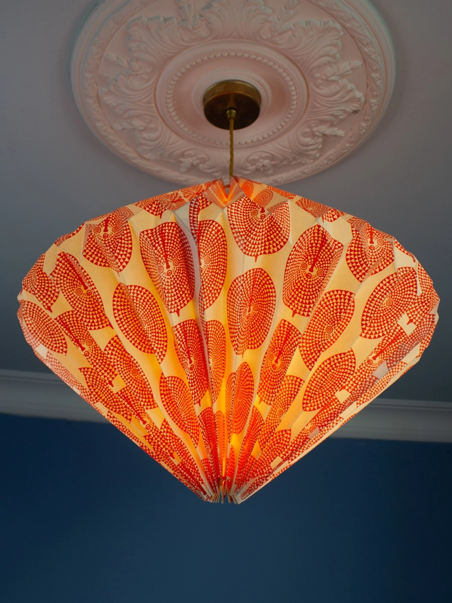 A pendant light with an orange and white geometric patterned shade hangs from an ornate ceiling rose against a blue wall.