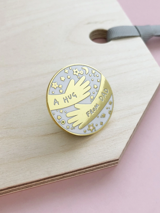 A white and gold pin badge with a hugging arms design and the words “A hug from Dad” is placed on a wooden tag.