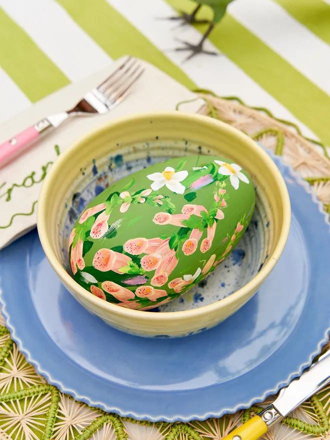 Soft green hand painted wooden egg as part of a wider tablescape, positioned in a yellow speckled bow on a blue scalloped plate. These are resting on a green jute tablemat atop a green and white striped tablecloth