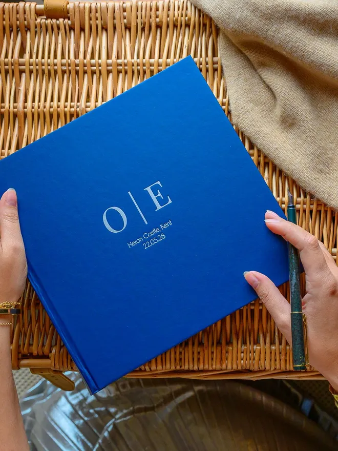 A blue hardback wedding guest book, printed with a couple's initials, rests on a wicker basket.