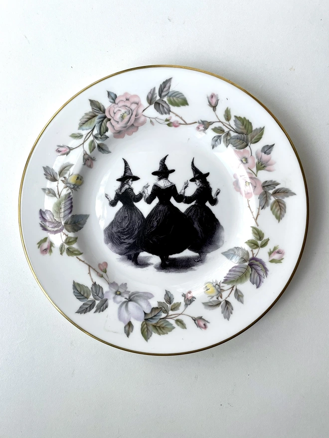 a coven of 3 witches dance hand in hand, the image is victorian in black and white and is printed directly onto an ornate vintage plate