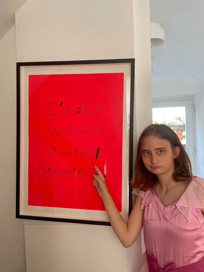 Piper pointing to her framed screen printed artwork which is her black font on a neon red background