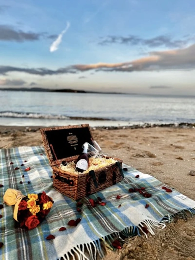 Stewart Muted Blue Picnic Blanket, a blue picnic blanket sat on the beach outside. It is accompanied by a wooden hamper of food and sits in front of the sea as a background. 