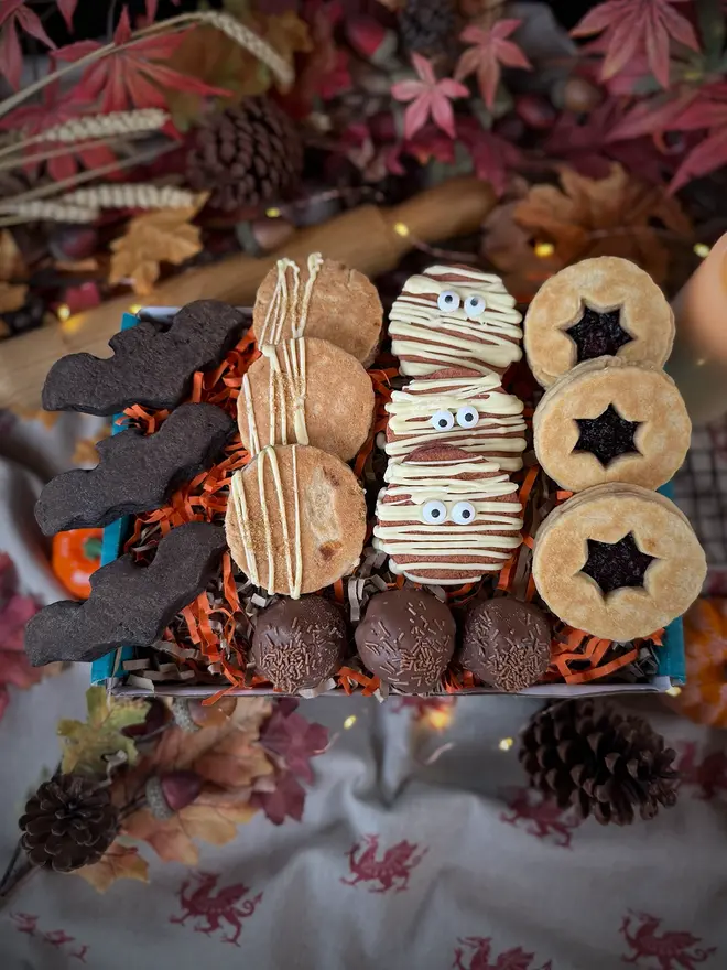 A Halloween-themed welshcake box filled with assorted treats, including red velvet mummy welshcakes, star-shaped blackberry sandwich welshcake, chocolate balls, and bat-shaped black velvet welshcake, and apple crumble welshcake arranged in orange shredded paper inside a blue box on a table with a candle and autumn leaves in the background.