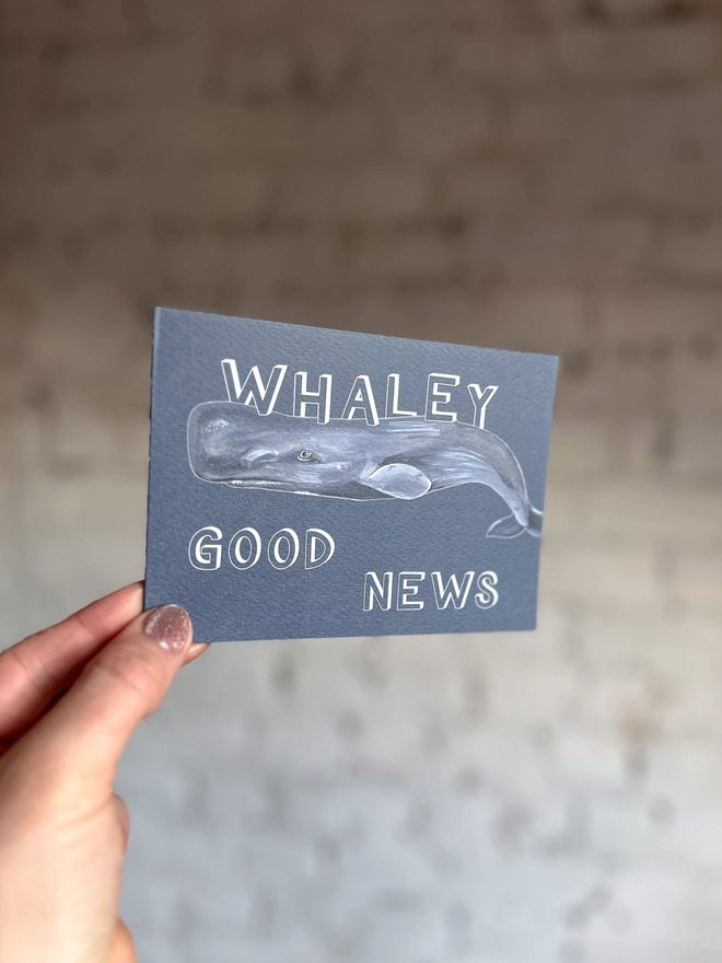 a dark grey blue greetings card featuring an illustration of a sperm whale with the text “whaley good news”
