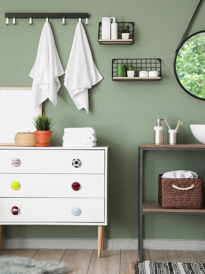 A set of six sports ball door knobs on a white chest of drawers with pine hair pin legs. The chest of drawers is in a modern bathroom with a sage green wall. There is a black vanity stand with brown wooden shelves and a modern shaped matt white sink. On the wall there is a black rimmed mirror, a row of hooks with white towels hanging down, a pile of white towels, black wire racks with white bathroom holders and a stainless-steel soap holder and toothbrush holder. The floor is wooden with a soft grey rug and a black and white rustic rug. The sports ball door knobs are made by Candy Queen Designs.