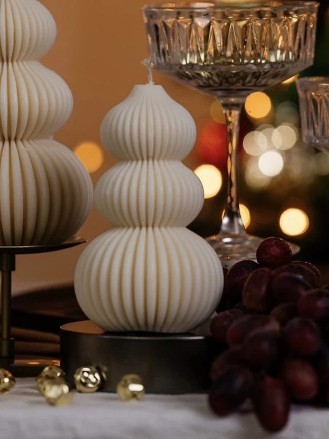 Signature Christmas Tree Sculpture Candle. A ribbed cream bauble-stack candle on a dark stand, styled with crystal coupe glasses, red grapes and gold bells. The textured sculptural design adds understated elegance to a Christmas drinks or dinner table.