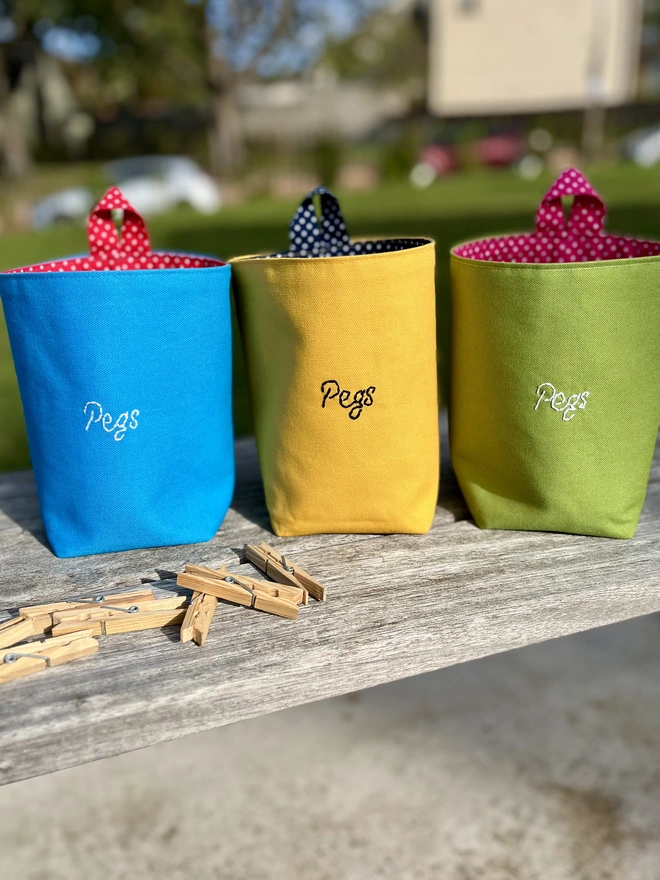 3 large cotton peg bags in bright colours, embroidered with 'Pegs' on the front, standing on a garden bench with clothes pegs scattered around.