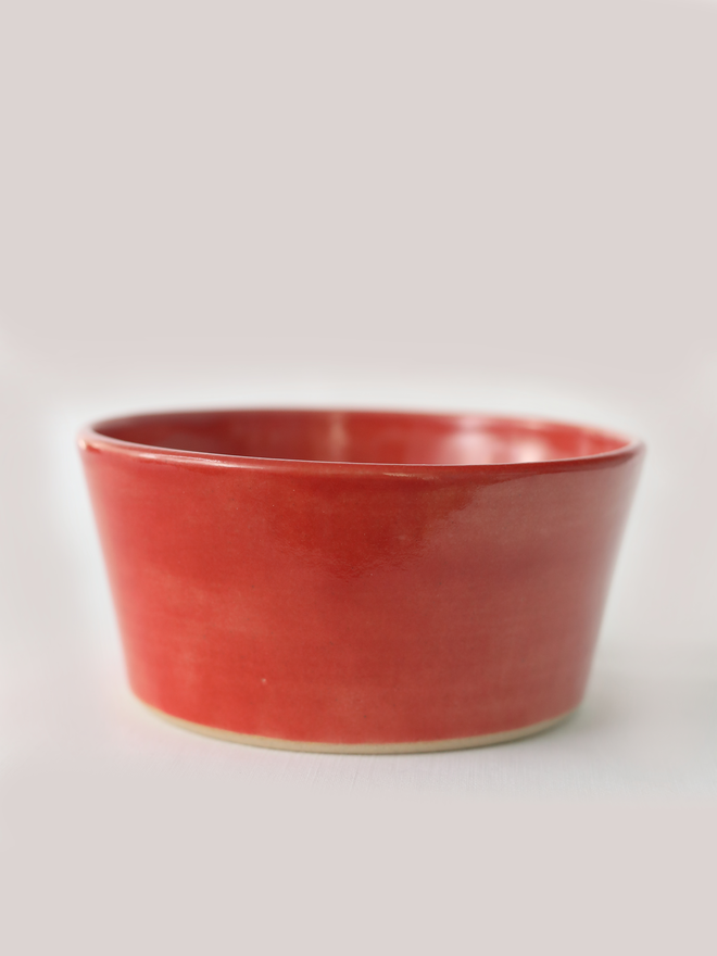 A single hand-thrown ceramic breakfast bowl in cherry red, with a glossy glaze showing subtle variation in tone, a gently flared form and unglazed natural clay base, photographed against a plain light grey background.