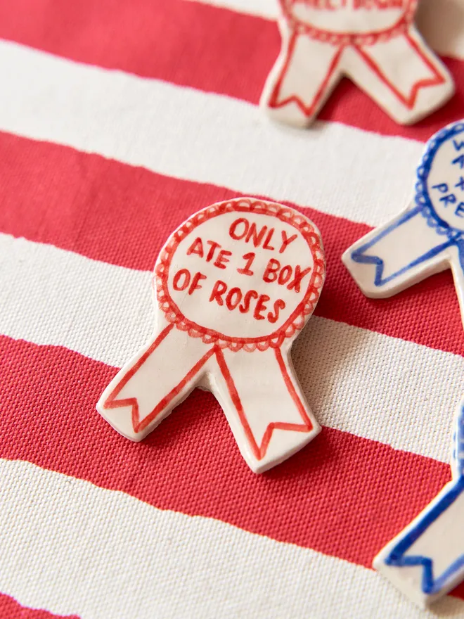 A selection of Red, Green & Blue Handmade Ceramic Christmas Award Badges, handpainted with different phrases, atop a striped red and white tablecloth