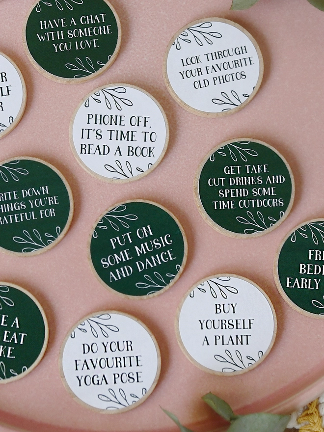 Several wooden tokens with dark green and white labels, each with a self care idea printed on, lay on a pink plate.