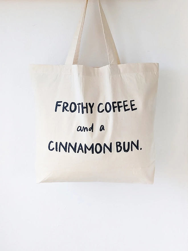 A natural coloured canvas tote bag with black slogan reading Frothy Coffee and a Cinnamon Bun.