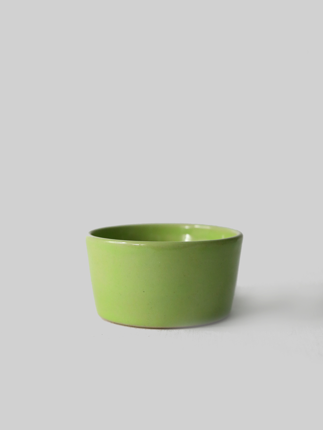 A small handmade ceramic side bowl in glossy spring green glaze, with a tapered sides and a wide open rim, photographed against a plain light grey background