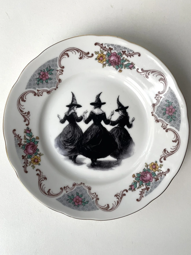 a coven of 3 witches dance hand in hand, the image is victorian in black and white and is printed directly onto an ornate vintage plate