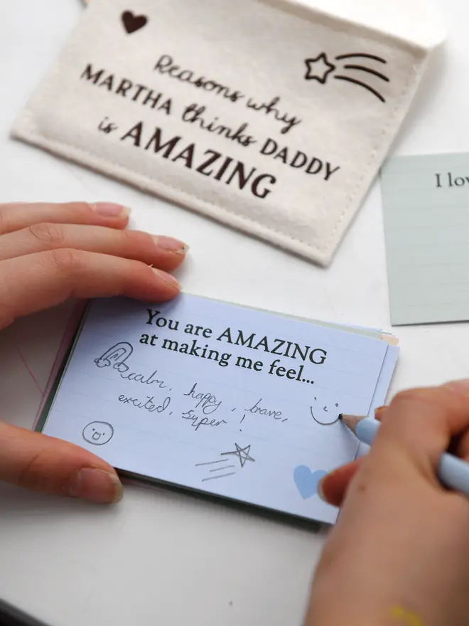 A child’s hands are shown writing on a light blue prompt card that says “You are AMAZING at making me feel…” using a green pencil. Other completed cards with handwritten messages are visible nearby on a white desk, including one that says “The best adventure we’ve had together was…” This scene captures a child filling out a sentimental Father’s Day gift.