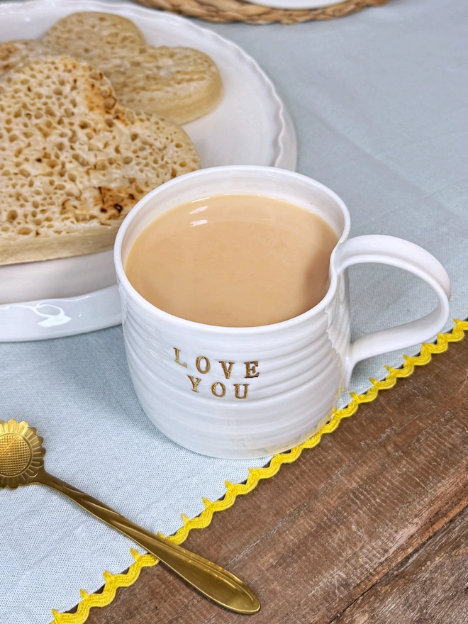 Love You hand thrown porcelain mug with gold lustre text on the side of the mug. The mug sits on a rustic wooden table top with some heart shaped crumpets on a plate and a pretty gold flower shaped tea spoon on a teal table cloth
