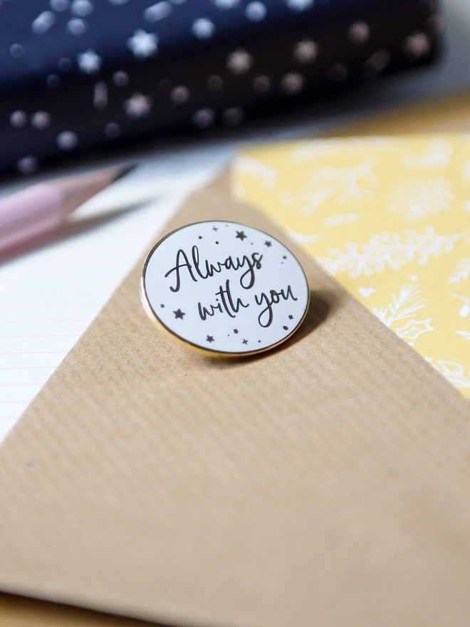 A white and gold enamel pin badge rests on a Kraft envelope. It has a subtle star design and the words "Always with you".