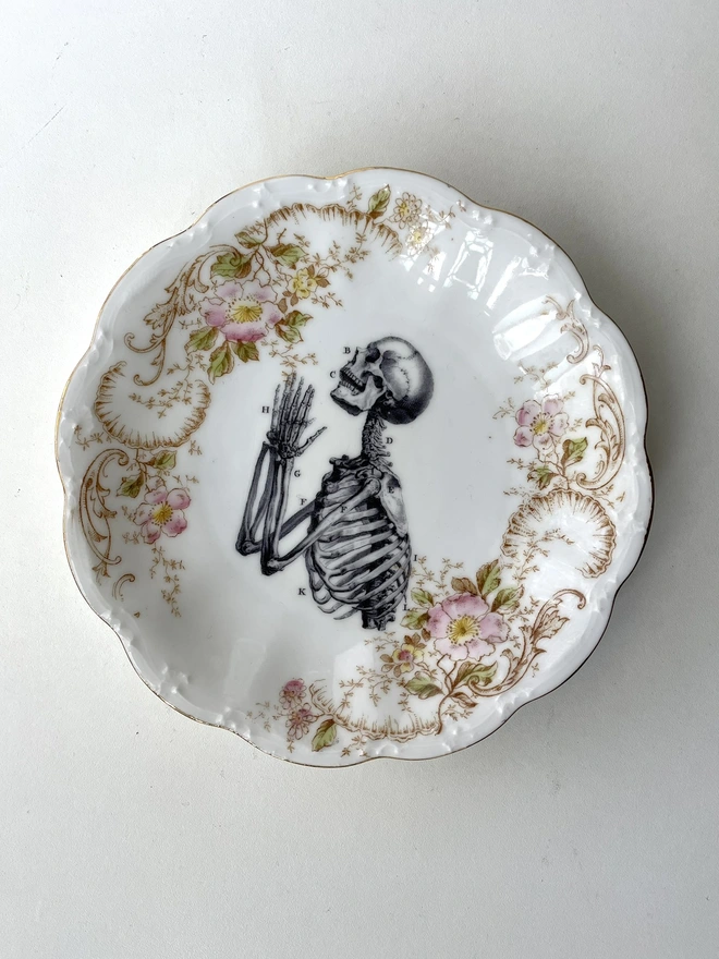 vintage plate with an ornate border, with a printed vintage illustration of a praying skeleton in the middle