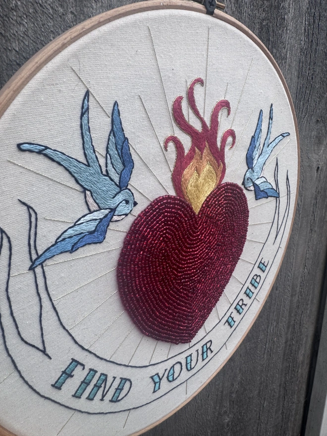 Embroidery hoop containing red beaded heart two blue swallows stitched fire and a banner containing find your tribe