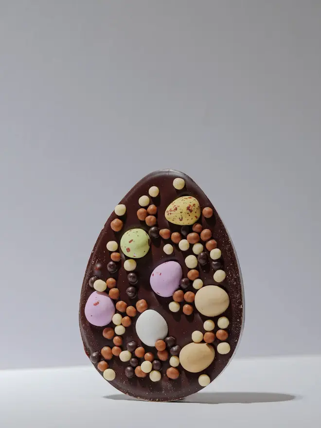 Large dark chocolate egg-shaped tablet decorated with pastel mini eggs and assorted chocolate pearls, standing upright on a plain white background.