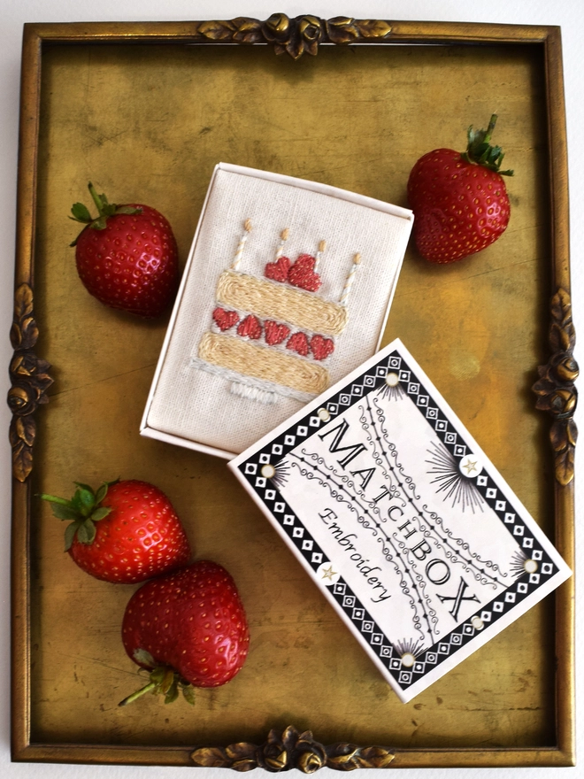 The finished embroidery placed inside the matchbox packaging for display.  Shown here on a vintage brass tray with scattered home grown strawberries.