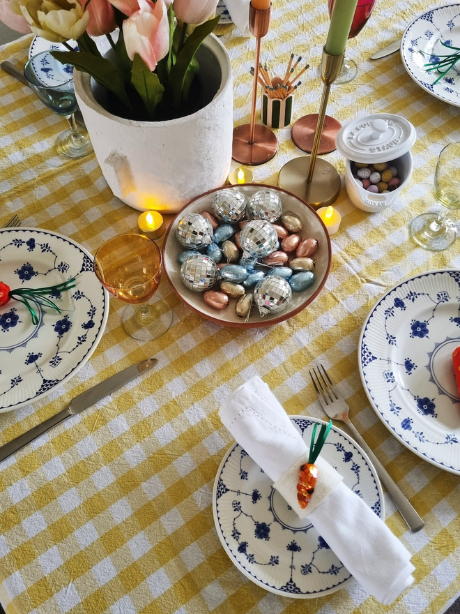 a table with yellow and white gingham cloth, blue and white plates, with a white napkin with sequinned carrot napkin ring around it