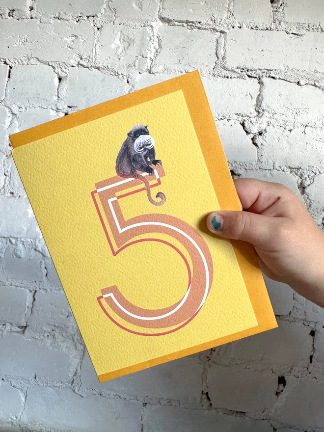 bright yellow card featuring a big orange number 5 and an illustration of an emperor tamarin with a bright yellow envelope