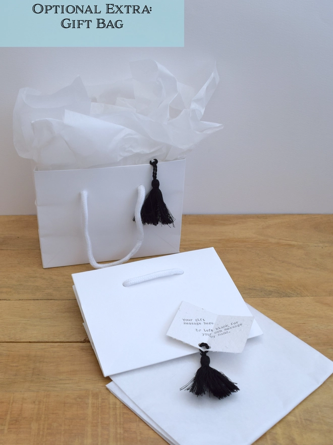 A white paper gift bag, approx. H120x W160x D80mm & rope handles, with white waxed tissue paper and a gift card embedded with seeds with a black tassel.