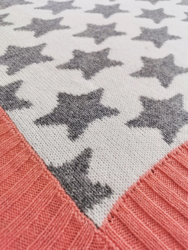  A close up of the corner of a knitted blanket showing the knit stitches, grey and white star design and coral pink ribbed trim.