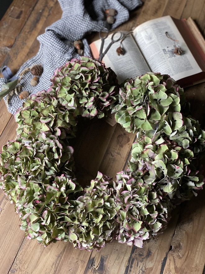 A dried green hydrangea wreath with burgundy accents, displayed on a wooden table with autumnal accessories.