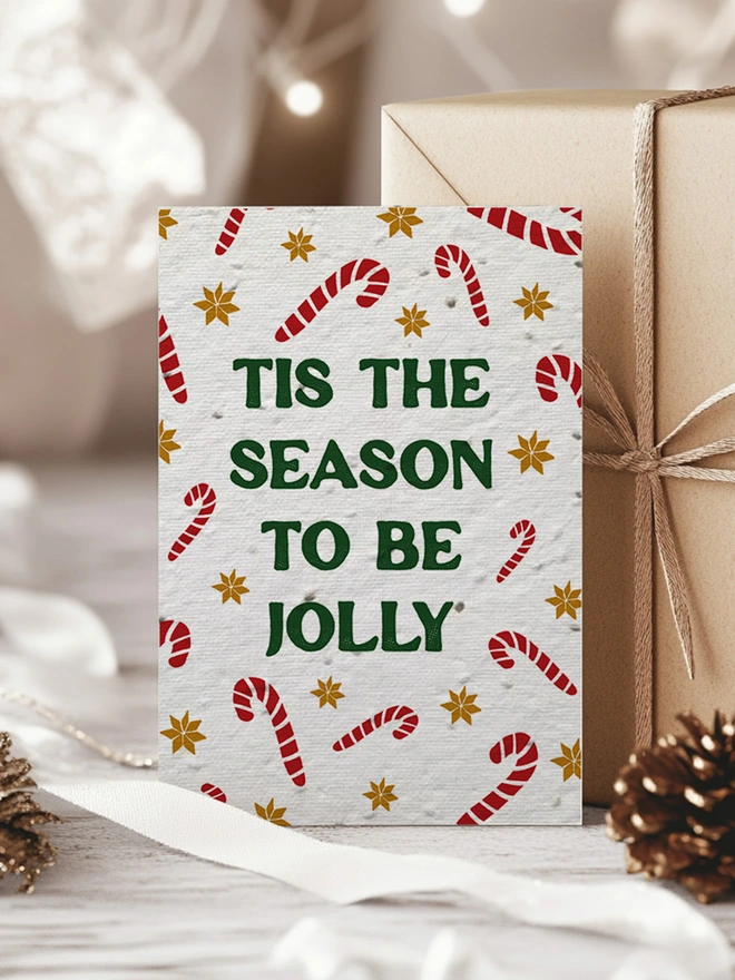 Christmas Card with candy canes and gold star pattern with 'Tis the season to be Jolly' in bold forest green text. The card is surrounded by Kraft paper wrapped presents and pine cones.