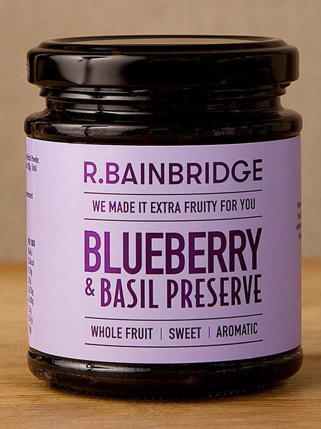 Blueberry Basil (Set Of 1, 2 Or 3), a jar of jam sitting on a wooden surface against a plain backdrop. 