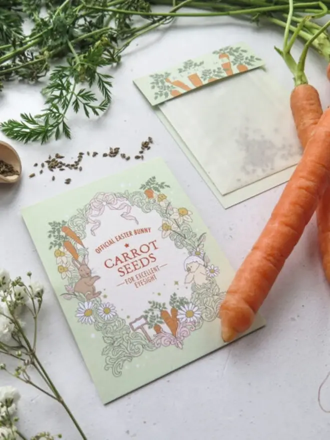 An angled view of the illustrated carrot seed card, with a fresh carrot resting on its corner. The "Plant Me" seed pouch and loose carrot seeds lie behind it.