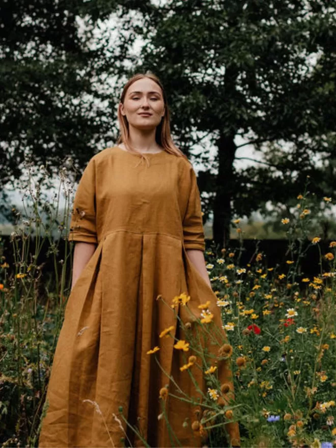 Midi length linen dress in golden colour. Grown on sleeve finishes at elbow. Deep pleated skirt.  Outdoor front view.