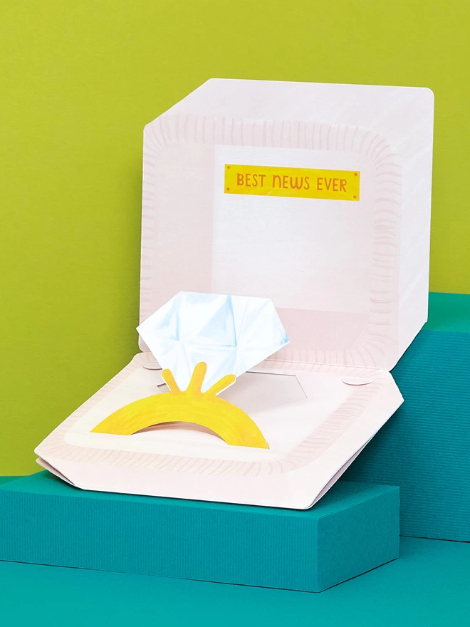 The 3D pop out card is set on a teal and green background. The pink engagement ring box with painted stitch detailing has a hand lettered ‘Best news ever’ message inside. Complete with pop out painted diamond ring 