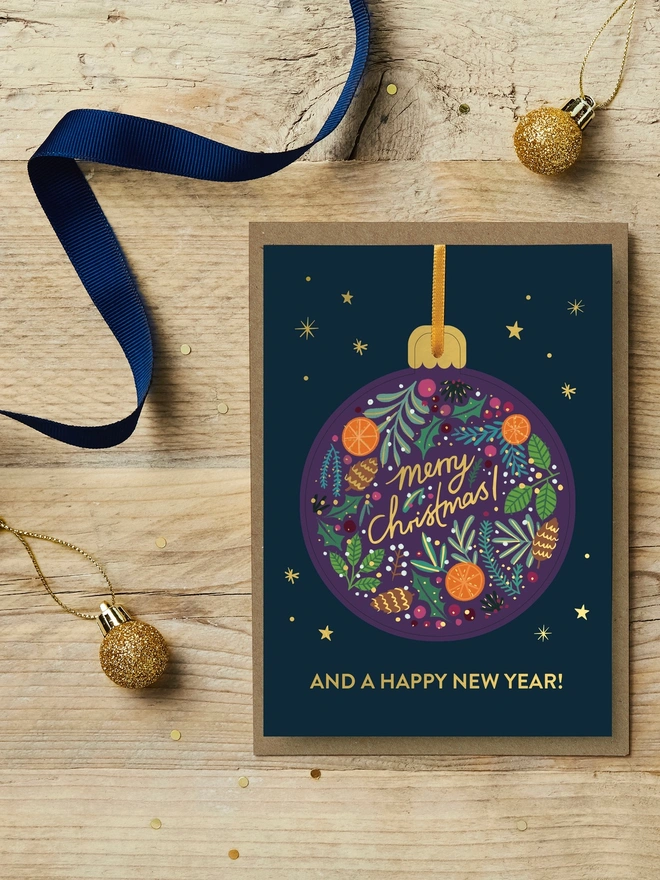 Christmas pop-out bauble card with festive foliage illustration on it sat on a wooden desk with blue and gold Christmas decorations.