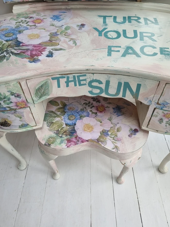 A painted dressing table in white and pale pink with large florals in blues and mauves and text in turquoise that reads 'turn your face to the sun'. The whole piece is distressed in the shabby chic style.