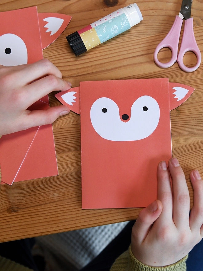 Several orange fox greetings cards lay on a wooden table. A child is gluing the ears to the corners of one card.