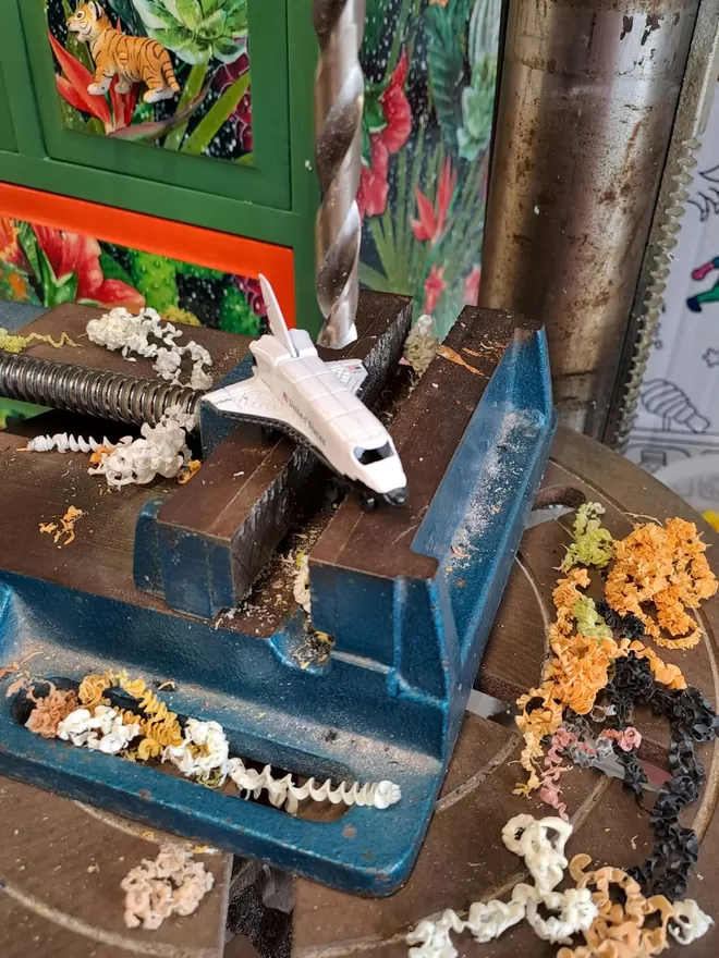 A small white metal and plastic toy space shuttle sits on a clamp on the table of a pillar drill. A chest of safari style drawers can be seen in the background. The space rocket is being drilled by Candy Queen Designs and made into a decorative space rocket dimmer switch for a child's nursery. 