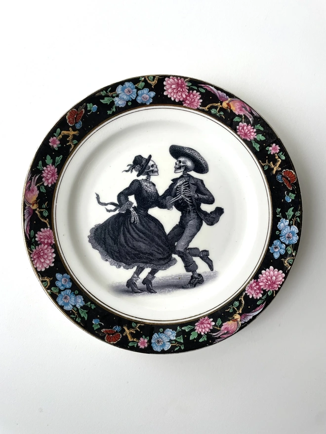 vintage plate with an ornate border, with a printed vintage illustration of two dancing day of the dead skeletons wearing a dress and a suit in the middle