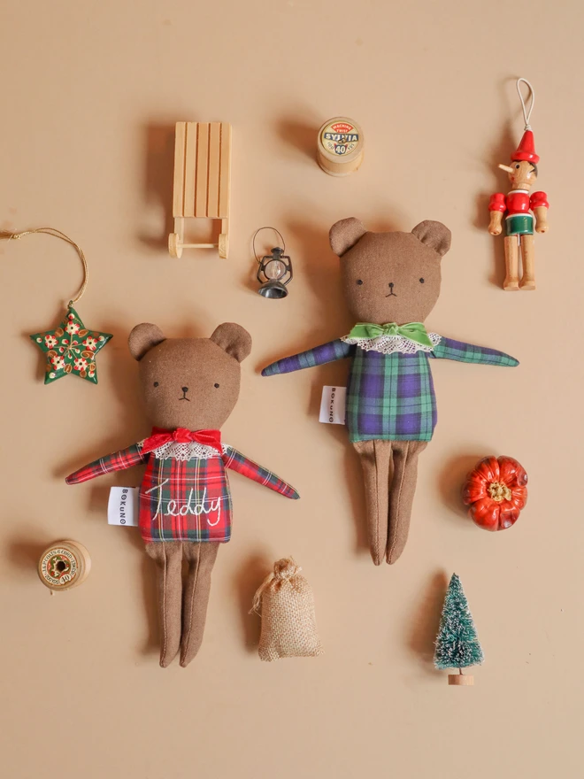 Two handmade linen teddy bears wearing festive tartan outfits, one in red and green with embroidered “Teddy” lettering and a red bow, and the other in green and navy tartan with a green bow. Both feature delicate lace collars and embroidered faces.