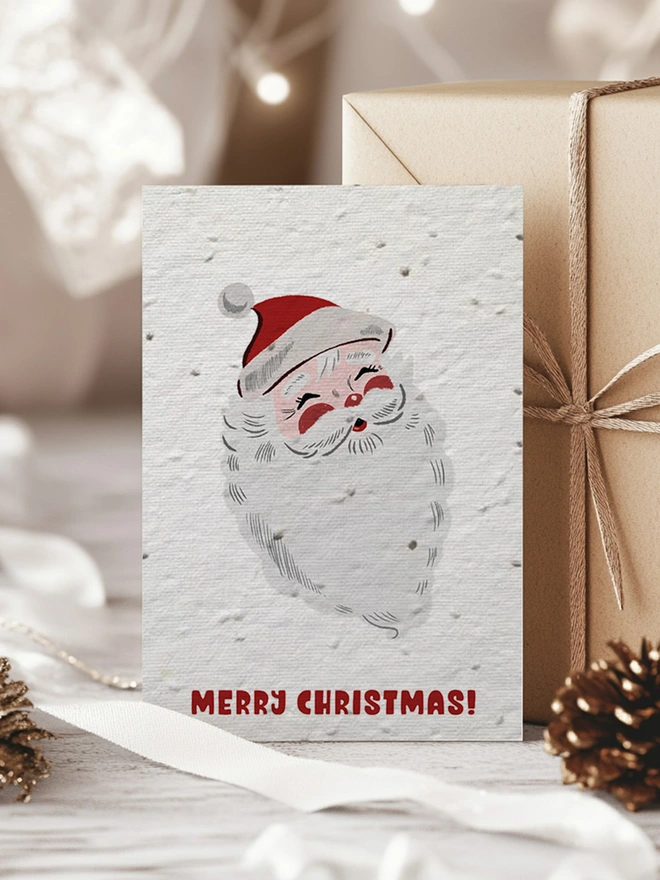 Christmas card with an illustrated Santa Clause with 'Merry Christmas' written underneath. In the background is presents in a Christmas scene