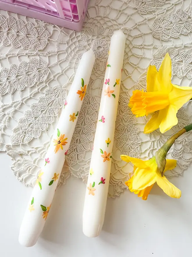 Blooming For You & Spring Bound Hand Painted Candle, a pair of taper candles against a white desk. The floral design can be seen wrapping around them. They are accompanied by a flower.