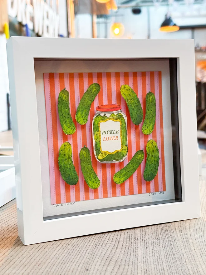 Charming framed collage featuring vibrant illustrated pickles, set against a colorful red and pink striped background, with gherkins and a pickle lover's jar. This vibrant and lovingly crafted mini collage is super cute and will delight pickle lovers and gherkin fans alike!