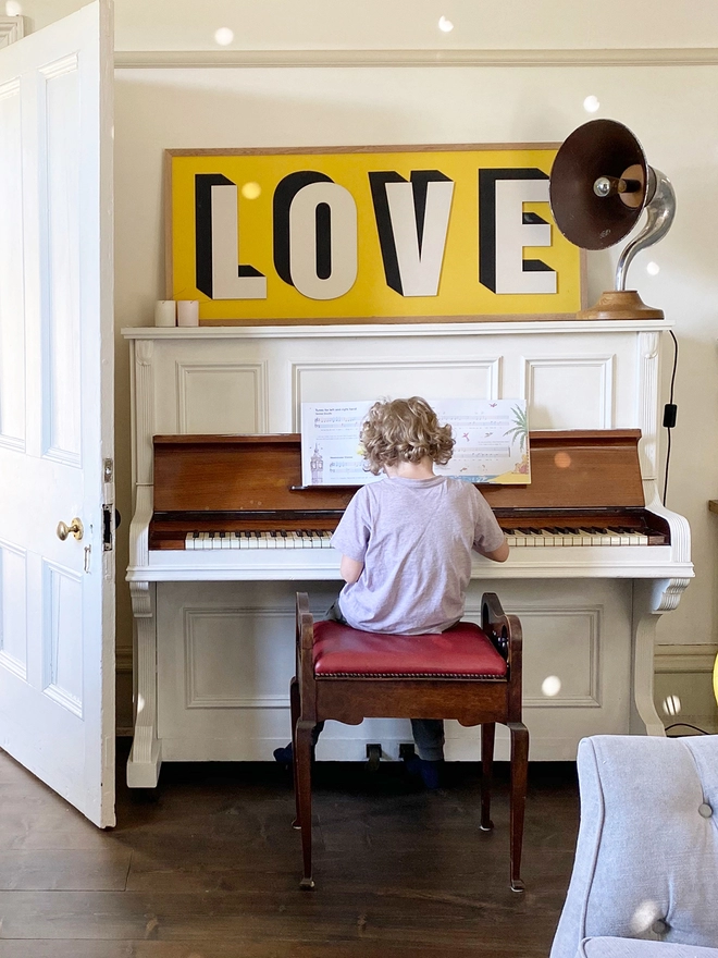 A child playing piano sat on a stool with a wooden painted sign resting above the piano reading LOVE on a yellow background