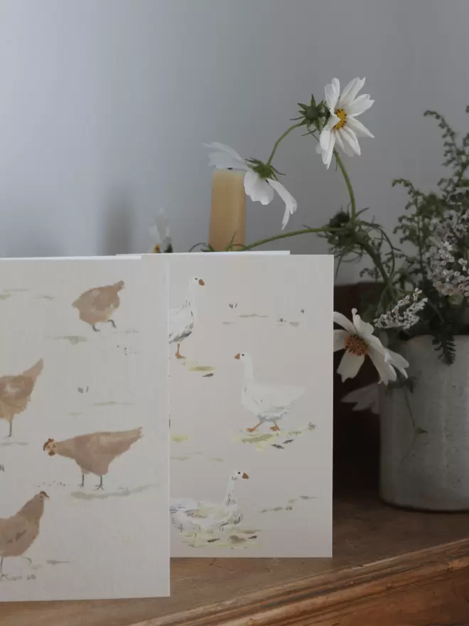 2 cards with chickens and geese on a wooden shelf next to a bud vase of cosmos. 