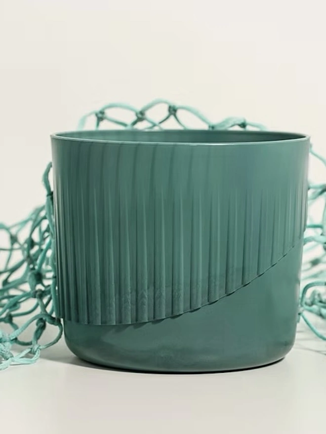 Ocean Plastic Plant Pot Small, a green recycled plant pot sitting on a white surface against a plain backdrop. It is accompanied by some recycled fishing nets.  