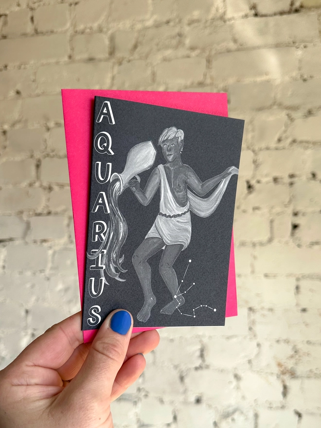 a greetings card featuring a greyscale illustration of a zodiac style water bearer with the text “AQUARIUS” with a neon pink envelope